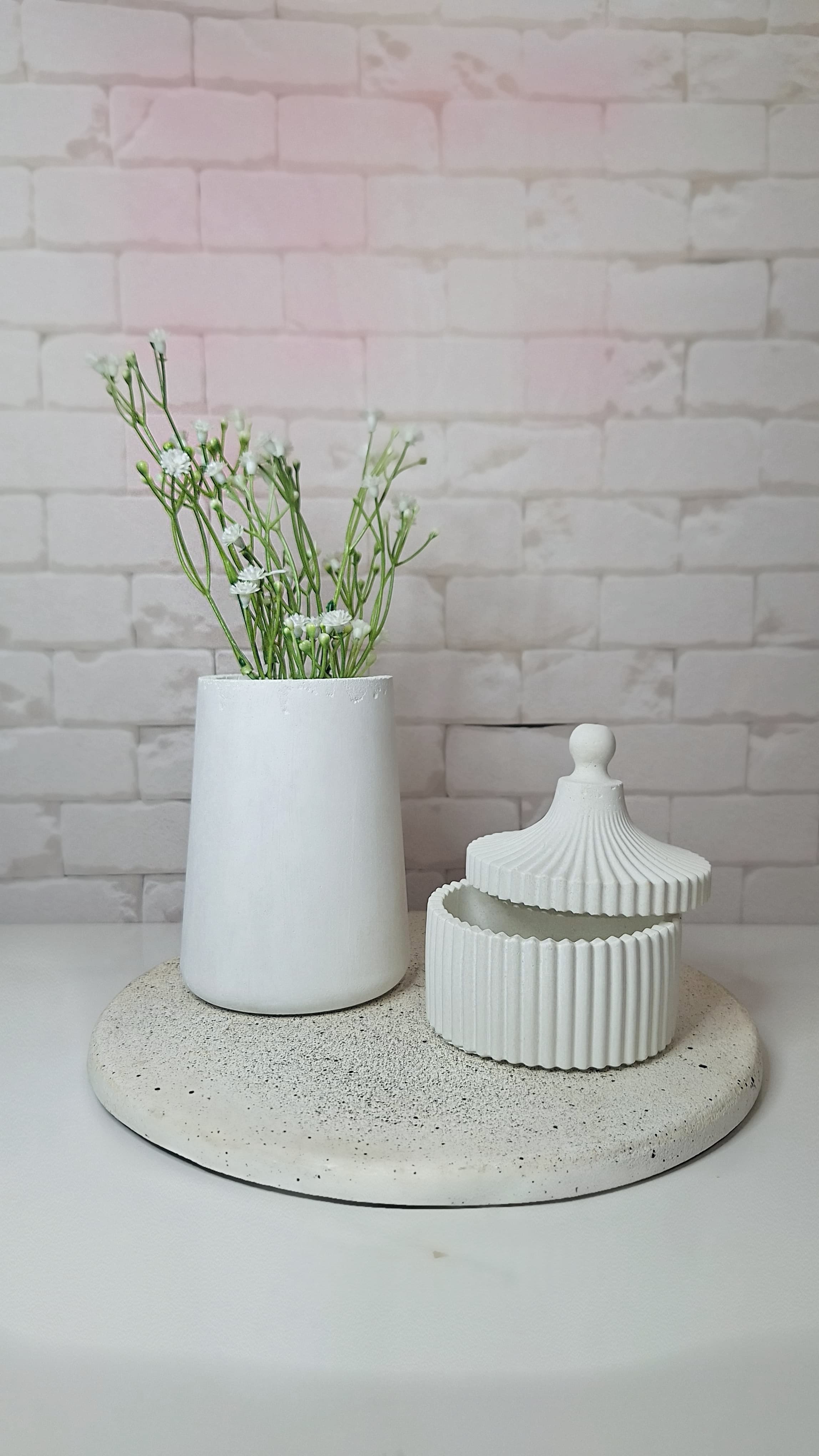 Imagem de 2 vasos de concreto brancos em cima de uma bandeja de concreto branca. O vaso maior possui uma planta dentro e outro menor contendo uma tampa