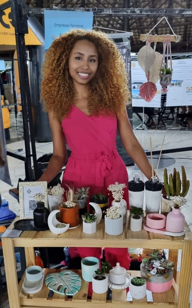 Imagem de uma mulher negra de cabelos cacheados, com uma camisa branca e que está de braços cruzados